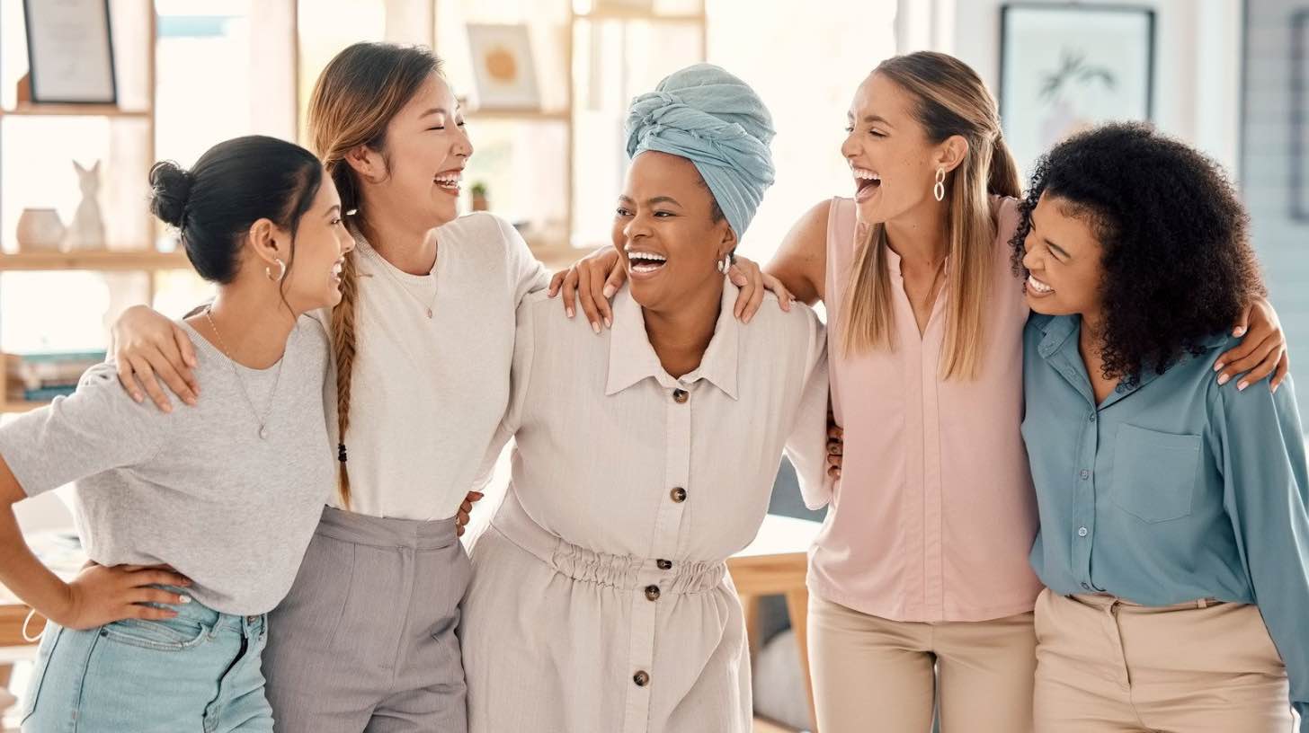 Eine diverse Gruppe von fünf Frauen steht eng zusammen und lacht gemeinsam. Sie tragen helle Business-Casual Kleidung in Pastelltönen und strahlen eine warme, einladende Community-Atmosphäre aus.